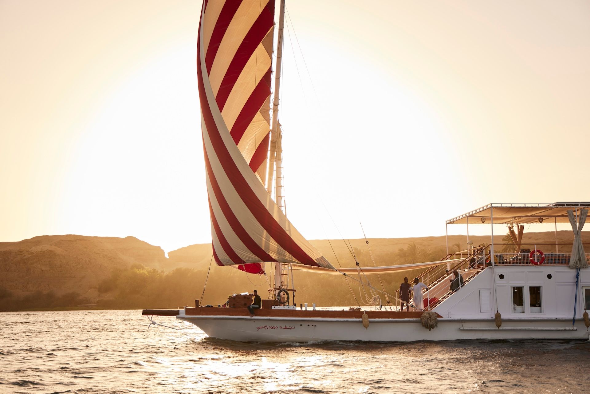 A Nour El Nil dahabiya at golden hour, red-and-cream striped sails against an amber sky on the Nile